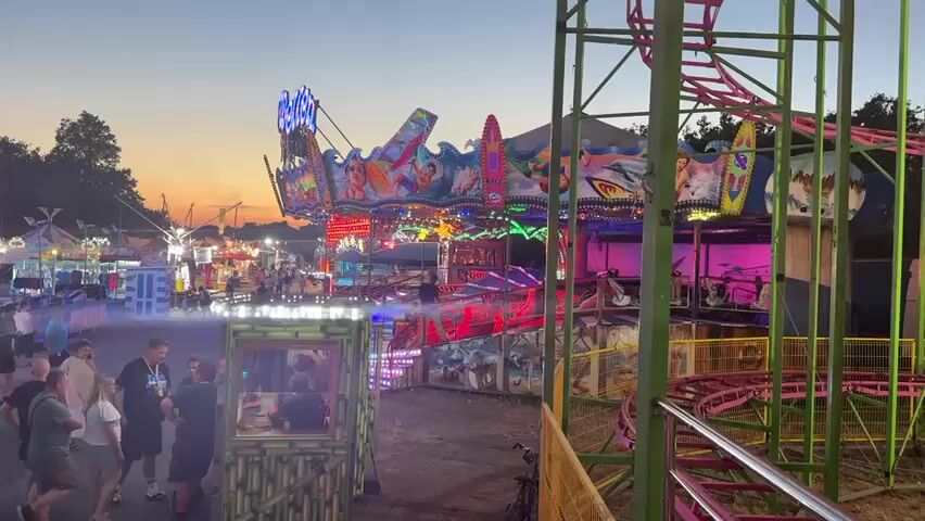 This media file depicts a lively amusement park scene at dusk, with a warm sunset casting a soft glow across the sky. The air is filled with the vibrant energy of a carnival, evidenced by the colorful lights of various rides and game stalls that illuminate the grounds.

In the foreground, a group of people are gathered near a small, bamboo-themed booth, some engaged in conversation while others appear to be observing the activities around them. Their postures suggest a relaxed and social atmosphere. Behind them, the dynamic amusement park unfolds. A prominent ride, with a brightly lit, oscillating platform featuring cartoonish characters and images of aquatic life, is in motion. Its flashing lights and colorful decorations create a festive and exciting visual. To the right, the structure of a roller coaster with its distinctive green and pink track is visible, hinting at thrilling rides to come.

Further into the park, more rides and attractions are visible, including what appears to be a smaller spinning ride and other game booths, all adorned with bright lights and signage. The overall scene suggests a popular event or fair, likely a weekend or evening gathering. The time of day, indicated by the sunset and the artificial lighting, adds a magical and atmospheric quality to the experience. The setting appears to be an outdoor carnival ground, possibly in a temporary location given the nature of amusement park setups. The mood is one of cheerful excitement and community enjoyment, characteristic of such events.