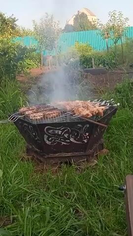 A person's hand holds a clear plastic bottle, pouring liquid onto skewers of meat cooking over a fire in a decorative metal brazier. The meat is arranged on two grill grates. Smoke rises from the cooking meat and the fire. The brazier sits on a base of what appears to be concrete blocks surrounded by green grass and foliage. In the background, a light-colored building and a green fence are visible. The sky is bright, suggesting daylight.