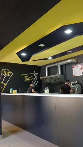 The video shows the interior of a fast-food establishment with a black and yellow color scheme. A long, dark counter is the focal point, with two employees in black shirts visible behind it. The ceiling features geometric yellow and black panels with recessed lighting. The walls are black, decorated with white chalk drawings of food items and a hand pointing. Various objects are on the counter, including drinks, jars, and kitchen equipment. The overall impression is of a modern, clean food service area.