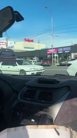 From the inside of a car, a view of a busy intersection is captured on a sunny day. The foreground is dominated by the car’s dashboard, covered with a gray, suede-like material. On top of the dashboard is a small device displaying a phone number: 87024908958.

Through the windshield, a bustling street scene unfolds. The camera is positioned at an intersection. A white Nissan Primera is stopped across the street, waiting for the light to change. To the right of the Nissan, there is a gray sedan. In the background, businesses line the street, including a "Sulpak" electronics store and a "TIM ROSE" flower shop. A pedestrian crosswalk with a crosswalk sign is visible near the flower shop.

The sky is a clear, bright blue, and the sun casts a warm light on the buildings and cars. Streetlights are spaced along the roadway.