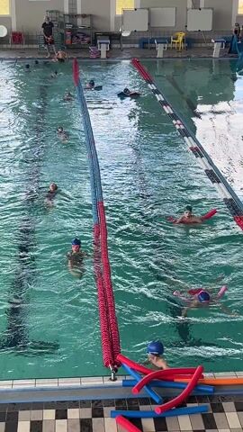 The video shows an indoor swimming pool. Swimmers, many wearing swimming caps, are spread throughout the pool in various lanes. Some swimmers use pool noodles for support, while others swim freely. A lifeguard appears to be supervising from the side of the pool. Lanes are marked with dividers. The pool is well-lit.