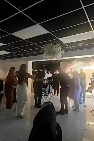 A group of people are dancing in a room with a checkered ceiling. A white floral decoration hangs from the ceiling. There are pink balloons and a lighted sign in the background. People are dressed in casual attire.