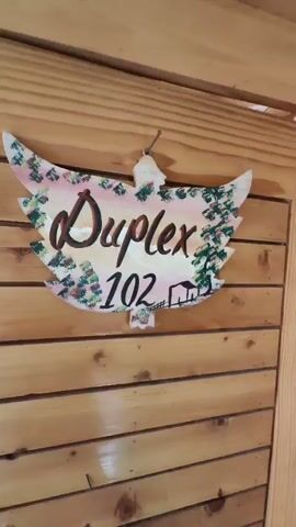 The video opens with a close-up of a hand-painted sign attached to a wooden wall. The sign is shaped like a leaf and reads "Duplex 102" in stylized lettering. The background of the sign features a colorful depiction of trees and a small house. The wall is made of horizontally aligned wooden planks. The camera then pans to the right, revealing the interior of a rustic wooden room. The room appears to be a duplex living space, with a bunk bed structure. The lower bunk is made up with white sheets, a patterned red and white blanket, and a small white towel. A decorative pillow with colorful designs sits at the head of the bed. Above the lower bunk, there is an upper sleeping area with a wooden railing. A blanket is folded on the upper bunk. A wooden ladder is positioned to the left of the lower bunk, providing access to the upper level. To the left of the ladder, a cream-colored curtain hangs next to a window, with sunlight filtering through. The walls and ceiling are constructed of unfinished wood, giving the space a cozy, cabin-like feel. The overall impression is of a simple, charming accommodation.