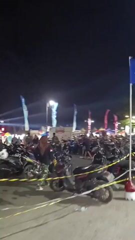 The video captures a nighttime scene from a slightly elevated perspective, looking across a densely packed area of parked motorcycles. Several flags are visible, including a prominent blue flag with an emblem on a tall pole in the foreground, and other flags further in the background. Bright beams of light are directed into the dark sky from the left side of the frame, suggesting some sort of event or performance is taking place. In the mid-ground, a crowd of people is visible milling around amongst tents and structures, indicating a public gathering or market. Yellow caution tape is strung across the lower portion of the frame, partially obscuring the motorcycles. The overall atmosphere appears lively and event-driven.