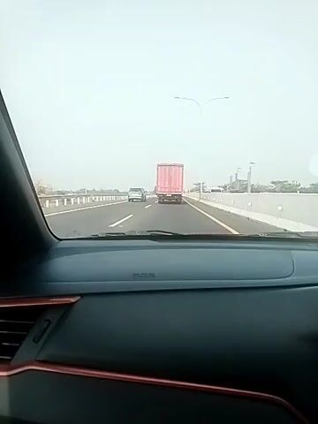 The video shows a view from inside a car driving on a highway. The driver is following a pink truck. There are other cars on the highway and trees in the background. The sky is overcast. The car's dashboard is visible in the foreground.