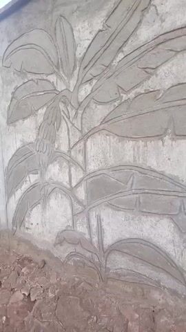 The video shows a close-up of a concrete wall with a relief sculpture of banana plants. The sculpture depicts a banana tree with large leaves and a bunch of bananas. The background is a rough, gray concrete surface, with some small rocks and debris at the bottom of the wall.  There is some blurred movement near the end of the video, but nothing clearly discernable.