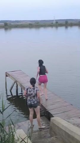 A person wearing a black vest and pink shorts jumps off a wooden pier into a body of water. Another person, wearing a patterned shirt and shorts, stands on the pier, facing away from the viewer. Two children, one in a pink hooded sweatshirt and polka dot leggings, and another in a black bikini top and pink shorts, stand on the shore, watching. The environment is a lake or river with a tree line in the background under an overcast sky.