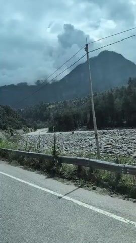 The video shows a road with a white line running down the middle, bordered by a metal guardrail. Beyond the guardrail, a wide, rocky riverbed is visible, with a small amount of water flowing through it. Lush green vegetation grows along the riverbank. In the background, a large, tree-covered mountain rises against a cloudy sky. Power lines are visible against the sky. The camera appears to be moving, suggesting the video was taken from a moving vehicle.