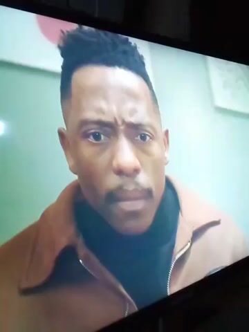 This video shows a close-up of a man's face with a concerned expression, followed by a shot of television credits with a sunset background and large lettering that appears to spell out "UZALO". The man in the first shot has dark skin, short dark hair styled in a high top fade, and a mustache. He is wearing a black turtleneck and a brown jacket. The second shot shows a list of names under the heading "CAST" against a colorful sunset.