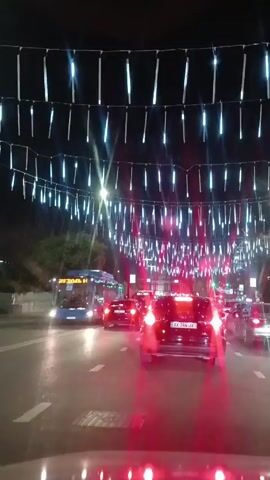 From the perspective of a driver in a vehicle, the video shows a street at night adorned with strings of bright, dangling lights spanning across the road.  Multiple vehicles, including cars and a bus, are driving on the wet asphalt street, their lights reflecting off the road.  The cars are in motion, and the general environment appears to be a city street with trees visible on the side. The lighting overhead is a prominent feature of the scene.
