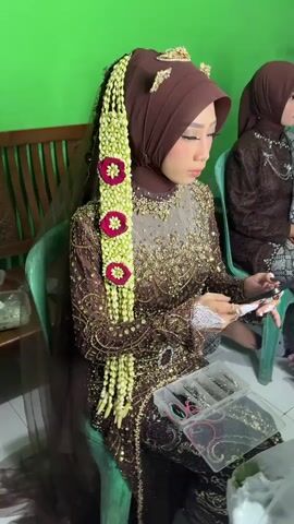 A woman is seated on a green plastic chair, wearing a brown hijab and a long brown dress adorned with gold embroidery and beading. Long garlands of white jasmine flowers with red floral accents are draped over her left shoulder. She is holding a small white object and touching her right index finger. In front of her, on an unseen surface, is a clear plastic organizer box containing colorful elastic bands and rings. The camera pans right, revealing a portable makeup case with multiple illuminated bulbs and a mirror, mounted on a tripod. The case holds various makeup items, a smartphone, and a white jasmine garland. To the right of the makeup case, another woman wearing a light-colored, possibly embroidered top, is seated on a green plastic chair, having her hair styled by an unseen individual. A man is visible standing in the background against a green wall. A woman wearing a patterned hijab briefly walks across the foreground, and another woman is kneeling on the floor.