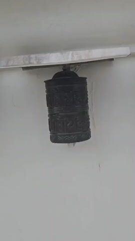A short video shows a row of prayer wheels mounted on a white wall. The prayer wheels are dark and cylindrical with embossed designs. They are attached to metal brackets under a metal overhang. The video pans from left to right, revealing three prayer wheels in succession. The lighting is dim, and the environment appears to be outdoors.
