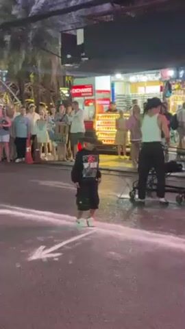 The video captures a dynamic street performance. A slender man with a white tank top and black trousers is dancing on a street. A child wearing a hat and a dark outfit joins him. They dance in front of an appreciative audience of diverse people. The performance happens on a street marked with a painted white arrow. The street scene is alive, with a booth selling goods and the soft glow of streetlights. Trolleys are parked next to the dancer. The background is filled with palm trees.