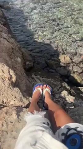 A first-person shot shows a person sitting on a rocky outcropping with their legs dangling over the edge. The person is wearing blue and white flip-flops, white pants with fringed hems, and a white top with a large blue and white circular design. Below their feet, clear water ripples over the rocks. Sunlight casts shadows on the rocks and the water.