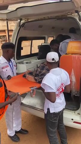 The video shows an ambulance with its rear doors open. Four individuals are visible. One person, wearing a white uniform with red trim and a black beret, stands on the left, holding an orange stretcher. Another person, wearing a white t-shirt with a red logo and a white cap, stands to the right, facing the ambulance and also holding the stretcher. Inside the ambulance, two other individuals, also wearing white, are partially visible. One is attending to a person lying on the stretcher, who is wearing a plaid shirt. The second person inside the ambulance is partially obscured. The scene appears to be outdoors, with reddish-brown ground visible. The ambulance is white, and some orange equipment, possibly medical supplies, is stacked inside. The individuals are engaged in the process of loading or unloading the stretcher and the patient into or out of the ambulance.