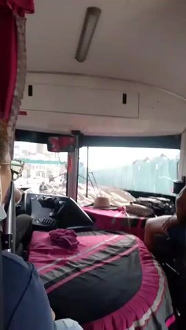 The video shows the interior of a bus. In the foreground, a person's shoulder and back are visible. In the mid-ground, a person's head and shoulder are visible on the left side of the screen, wearing glasses and a light-colored mask. The driver's seat is covered with a pink and black patterned comforter. A pink cloth is draped over the steering wheel. A straw hat and other items are visible on the dashboard. A person's head and shoulder are visible on the right side of the screen. Through the windshield, construction scaffolding and fencing can be seen. The environment appears to be a city street.