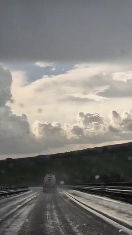 The video shows a view from inside a vehicle traveling on a wet highway. Raindrops are visible on the windshield. A large tanker truck with a white cylindrical tank is ahead on the right lane of the highway. The truck has a logo on the side. The highway is wet and reflective, indicating recent rainfall. The sky is cloudy with dramatic cumulonimbus clouds, and there are patches of lighter sky. Green hills are visible in the background, beyond the highway. The overall impression is that of driving in challenging weather conditions.