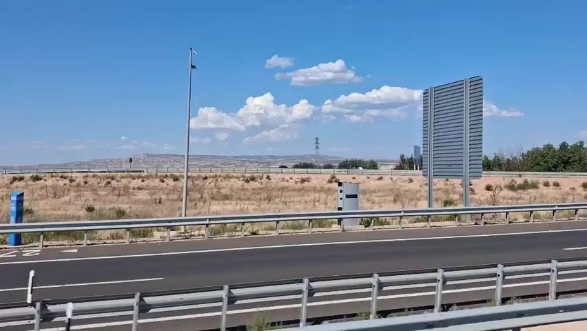 A highway with a clear blue sky and scattered white clouds. The road is empty and appears to be a highway with a metal guardrail on the side. Vegetation is visible on the sides of the road, and there are some structures in the distance, including what looks like a power line tower. The scene is captured from a moving vehicle, likely a car or bus, as indicated by the slight motion blur and the perspective.