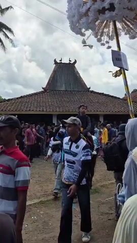 A group of people are gathered outdoors, with a traditional building in the background. Several horses are present, adorned with colorful blankets and decorations. Some individuals are riding the horses, while others are walking alongside them or observing the scene. One person is dressed in black and riding a horse, while another person in a blue dress with a veil is also on horseback. The atmosphere appears festive, with a crowd of onlookers present. The ground is dirt, and there are trees and palm trees visible. The sky is partly cloudy.