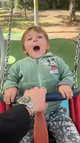 A young boy with short brown hair, wearing a green hooded sweatshirt and patterned pants, sits in a baby swing at a playground. The swing is made of metal chains and has a red and blue plastic seat with a crossbar. The playground has artificial green turf with circles of different colors on it. An adult’s hand with a silver ring and watch appears in the foreground, pushing the swing. The adult says "Woosh." The child laughs and seems to be enjoying the swinging motion. The camera angle changes slightly to show more of the surrounding environment, including trees and a yellow and white pole.
