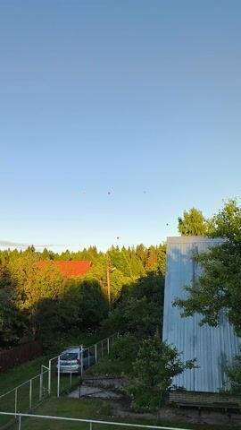 The video shows a bright, clear sky with a few balloons floating upwards. The balloons are small and colorful, appearing as dots against the blue. In the background, a line of trees forms a dark green horizon. To the right, a silver car is parked in a lot next to a building with a corrugated metal exterior. A red-tiled roof is visible amongst the trees. The sun appears to be setting or rising, casting a warm glow on the scene. The overall mood is peaceful and serene. There are no people visible in the video. The time of day appears to be late afternoon or early evening, given the angle of the sun and the soft light. The location is likely rural or suburban, given the presence of trees and open space. No text is visible in the video.