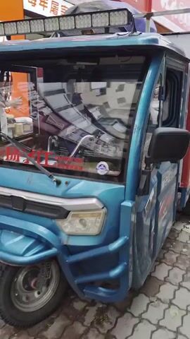 The video begins with a close-up shot of a blue and red three-wheeled delivery vehicle. The vehicle has a cargo box on the back with the J&T logo and a cartoon rabbit image. The vehicle is parked on a patterned brick sidewalk in front of a building. The camera then pans to the right, showing a woman in a yellow plaid shirt and gray pants walking away from the vehicle. She is carrying a white bag. The camera continues to pan, revealing more of the building's storefronts and then a row of parked cars. The scene appears to be an urban or suburban environment.