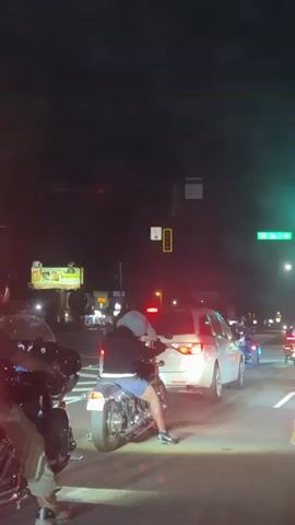 The video depicts a night scene at a traffic intersection where a group of motorcyclists are gathered. The motorcyclists are waiting at a traffic light that is green, indicating they have the right of way. A white minivan is positioned in front of the motorcycles, also waiting. The surroundings are dark, illuminated by streetlights and the lights of the vehicles. A billboard is visible in the background, and a street sign.