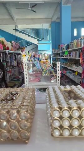 The video provides a close-up, eye-level view of two decorative tissue boxes on a white surface, likely a counter or table, in what appears to be a retail store. The tissue box in the foreground, slightly to the right, is adorned with a dense pattern of small, round, white faux pearls arranged in a grid. The tissue box on the left, partially obscured by the one on the right, is decorated with larger, clear, multifaceted faux gems, also arranged in a diamond pattern. Both boxes have a gold-toned base structure. The background of the video reveals the interior of a store. On the left, shelves are stocked with various items, including what look like packaged goods, crafts, and perhaps stationery. There are also some artificial flowers displayed. In the center, a white metal shelving unit holds an assortment of colorful items, possibly pens, art supplies, or small decorative objects, including some pastel-colored umbrellas or parasols. Further back, a blue staircase with white railings ascends, suggesting a second level or mezzanine. To the right, more shelves are visible, lined with numerous products, including containers and various retail items. The overall lighting suggests an indoor environment, possibly with natural light supplemented by artificial lighting. The store appears to be well-stocked and organized.