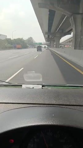 The video is captured from the perspective of someone driving a vehicle on a highway. The driver's dashboard and steering wheel are visible in the foreground. The road ahead is paved, with multiple lanes marked by white and yellow lines. An elevated highway structure runs parallel to the current road, supported by concrete pillars. Several vehicles, including cars and trucks, are traveling in the same direction. The sky appears hazy or overcast, suggesting it might be daytime. The scene is characterized by the movement of vehicles on a busy highway under an overpass.