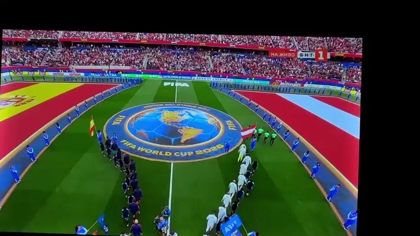 This is a video clip of the opening ceremony of the FIFA World Cup 2026, taking place on a lush green football field within a stadium packed with spectators. The scene is set at dusk, with the stadium lights illuminating the field.

The central focus is a large, circular mat featuring a globe illustration with the text "FIFA WORLD CUP 2026" around it. Two lines of people, presumably the national teams, are entering the field from opposite sides, forming an aisle. One team is dressed in dark blue uniforms, while the other wears white. They are flanked by individuals in blue uniforms holding flags and lining the pathway.

Large national flags are displayed prominently along the sidelines of the field, with the Spanish flag on the left and what appears to be the Austrian flag on the right. The spectators in the stands are a blur of red, suggesting a lively and enthusiastic crowd.

Visible text includes "FIFA WORLD CUP 2026" on the central mat. In the upper right corner of the screen, there is Russian text that appears to read "НА ЖИВО" which translates to "LIVE", and a channel logo with the number "1". Other sponsor logos, such as "BANK OF AMERICA" and "Hisense", are visible in the background along the stadium's advertising boards. The overall atmosphere is one of anticipation and excitement for the upcoming tournament.
