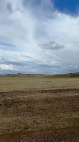 This media file depicts a vast, open grassland landscape under a dramatic sky. The scene is dominated by rolling hills covered in dry, golden-brown vegetation. In the foreground, a dirt road or track winds through the terrain, its surface appearing somewhat uneven and speckled with small stones. The horizon is marked by the undulating line of the hills, with a few rocky outcrops visible on the crest of the furthest ones.

The sky is a prominent feature, filled with a mixture of bright blue patches and large, dense white and gray clouds. The lighting suggests it could be late afternoon or early evening, with the clouds potentially indicating approaching weather. The overall impression is one of a remote, natural environment.

There are no people or specific objects clearly identifiable in the frame, emphasizing the natural and expansive quality of the setting. The activity, if any, is implied to be travel or movement along the dirt track through this sparsely vegetated landscape. The mood evoked is one of solitude and the raw beauty of nature. The location context provided is Bakty, Kazakhstan, which aligns with the visual cues of a steppe or grassland environment. There is no visible text in the media file.