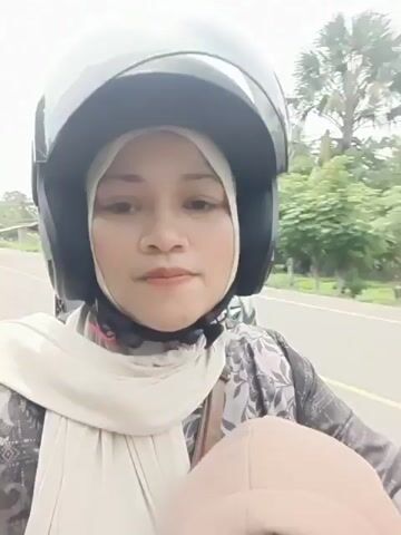 A woman wearing a hijab and a black helmet is on a motorcycle. She is looking at the camera and smiling. Behind her, a child wearing a black baseball cap and a hooded sweatshirt is partially visible. They are on a road with trees and palm trees in the background. A car is visible behind the woman.