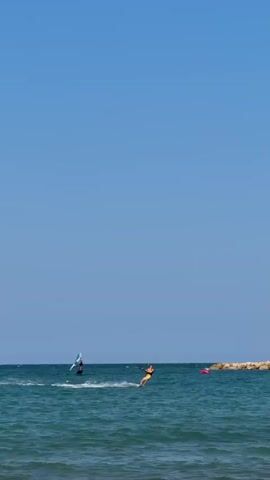 The video shows a clear blue sky over a tranquil ocean. Two individuals are engaged in water sports. On the left, a person is windsurfing with a blue sail. To the right of the windsurfer, a kiteboarder is captured mid-action, leaning back as they are pulled across the water by a kite. The water is a deep blue, with small waves and white spray trailing the kiteboarder. In the background, a man-made jetty constructed of large rocks stretches into the sea, with a few small yellow buoys visible near its end. The scene conveys a sense of leisure and athletic activity on a sunny day.