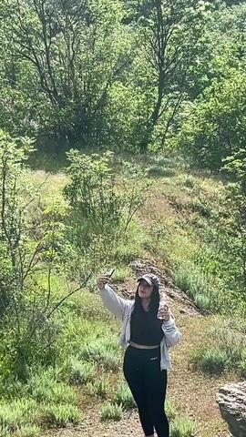 A woman is taking a selfie in a mountainous area. She is wearing black leggings, a black crop top, a light-colored jacket, a hat, and pink sneakers. She is standing on a rocky outcrop overlooking a valley filled with green trees and hills. The sky is bright with some clouds and the sun is shining brightly. The woman extends her arm towards the landscape.