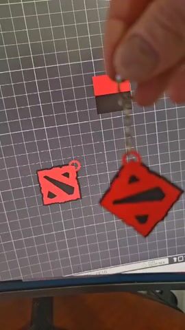 A hand holds a keychain with a red and black diamond-shaped pendant featuring a stylized "D" symbol, reminiscent of the Dota 2 logo. Another identical pendant lies on a gray, grid-lined surface, possibly a cutting mat. A small red rectangular object is also visible in the background on the same surface. The keychain is being manipulated, causing the pendant to swing and reveal its black backing. The lighting suggests an indoor setting, and no specific time of day or weather conditions are discernible. There is no visible text in the media. The overall mood is neutral, focusing on the display of the object.
