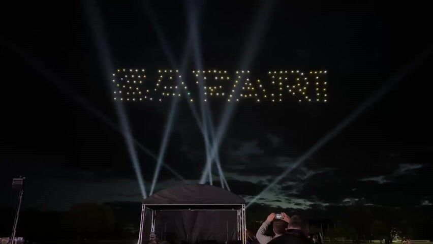 The video shows a nighttime scene of a drone light show. In the sky, a formation of drones creates the word "ROTARI." The drones are illuminated in yellow and blue. Beams of light shine upwards behind the word. Below, there is a small, covered stage with people standing in front of it, observing the show. One person is holding up a phone to take a picture or video. Trees are visible in the background under a cloudy sky. The lighting suggests a planned event or performance. The audio includes music.