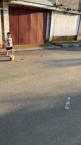 A young boy in a white t-shirt and black shorts stands on an asphalt street, facing away from a building with a brown garage door and a white wall. A red, white, and black soccer ball is positioned in front of him. A clear plastic water bottle stands upright further down the street to the right. The boy kicks the soccer ball with his right foot. The ball travels forward, strikes the standing water bottle, and knocks it over. The boy watches the bottle fall and then looks towards the camera. The scene is outdoors in daylight, with shadows cast on the asphalt.