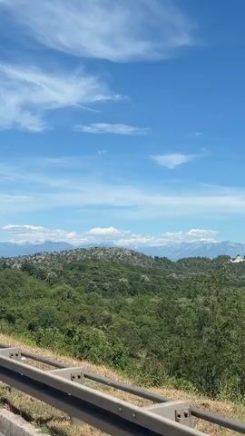 The video shows a scenic view of green hills and mountains under a blue sky with scattered clouds. A guardrail is visible in the foreground, suggesting the footage was taken from a vehicle moving along a road. The landscape is characterized by lush vegetation and a hazy mountain range in the distance. There are no people or animals visible in the frame. The overall impression is one of a peaceful, natural environment.