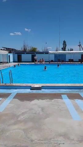 A wide shot captures an outdoor swimming pool on a clear, sunny day. The pool is filled with bright blue water and has a light blue border. Several people are in the pool, some swimming, others standing or sitting in the shallow end. A concrete deck surrounds the pool, with some blue painted lines visible on the surface. In the background, a row of buildings with white awnings runs along the side of the pool. Further back, trees and mountains are visible under a blue sky with scattered clouds. Some people are sitting on lounge chairs under the awnings. The overall atmosphere appears relaxed and recreational.