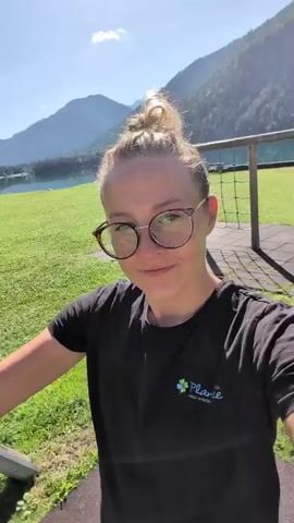 A woman with blonde hair pulled back and wearing glasses and a black t-shirt with a logo is on a swing set. She is filmed from a first-person perspective as she swings back and forth. In the background, there is a grassy field, a body of water, and mountains under a clear blue sky. She smiles at the camera and sticks her tongue out at one point.
