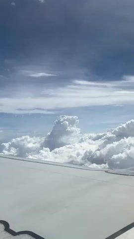 The video shows a view from an airplane window. The wing of the plane is visible in the foreground, and beyond it, there are layers of fluffy white clouds against a blue sky. The clouds vary in size and density, creating a textured landscape.  The light and sky create a soft contrast.