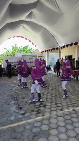 A group of young women in purple and white marching band uniforms, including hijab-wearing performers, are dancing outdoors under a white tent. Some are holding batons, while others appear to be playing drums. They are positioned on a patterned concrete surface. In the background, there are people watching, some seated, and several men holding drums and drumsticks. There are also banners and signs in the background, one of which appears to display text in Indonesian. The environment is lit by daylight, and there are green plants visible in the distance.