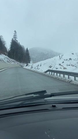 The video depicts a view from inside a car driving on a wet, winding road in a snowy, mountainous environment. The windshield wipers are actively clearing moisture. The roadside is lined with snow and guardrails, with trees visible in the background. The sky is overcast, and the visibility appears limited. There is a small structure with a parking sign visible on the right side of the road. No people are clearly visible.