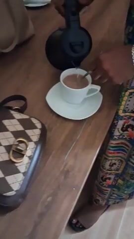 A person is preparing a drink on a wooden table. They are pouring water from a black electric kettle into a white cup that is sitting on a white saucer. The cup appears to contain a brown beverage, and the person is stirring it with a small spoon. A diamond-patterned handbag with a large gold buckle is on the table next to the saucer. The person is wearing a colorful patterned skirt and silver sandals. Other cups and a light-colored purse are visible in the background.