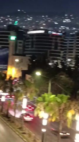 The video shows an aerial view of a city at night. In the foreground, there are palm trees and streetlights. Beyond the trees, there are several buildings, some of which are illuminated. In the distance, the city lights stretch as far as the eye can see. The sky is dark, with a few visible stars. There is no significant movement or action depicted in the video, other than the slight camera shake.
