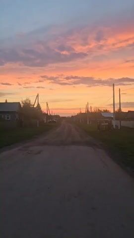 The video depicts a rural dirt road running through a small village at sunset. The sky is ablaze with vibrant shades of pink, orange, and yellow, interspersed with light, wispy clouds.  Wooden utility poles line both sides of the road, carrying power lines overhead.  Simple, single-story houses with fenced yards are visible along the road.  The road itself is gray and uneven, with patches of grass growing alongside it. The camera appears to be stationary, providing a fixed perspective of the scene.  No people or animals are visible.  The overall setting is peaceful and quiet, capturing the tranquility of a rural landscape at dusk.