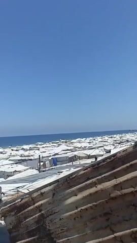 This media file presents an aerial view of a densely populated area with numerous temporary structures, likely tents or makeshift shelters, clustered together. The structures are predominantly white, with some featuring blue tarps. They are situated on what appears to be a sandy expanse, possibly a beach, with the ocean visible in the background. The water is a deep blue, and the sky is clear and bright blue, suggesting a sunny day.

In the foreground, a dilapidated, rusty metal structure, possibly a roof or part of a building, dominates the lower portion of the frame. A vertical, weathered metal pipe or pole stands prominently in the mid-ground, partially obscuring some of the shelters. There are a few individuals visible in the distance, scattered among the shelters, indicating human presence and activity within the settlement. The overall impression is one of a crowded, informal settlement, potentially a refugee camp or temporary housing area, situated close to the sea under clear, sunny weather. The scene evokes a sense of resilience and perhaps hardship, given the rudimentary nature of the structures. No visible text is present in the media file.