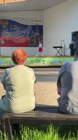 Two people are seated on a wooden bench, their backs to the camera, watching a young girl perform on a stage. The girl is wearing a white top and a red skirt, holding a microphone. Behind her, a banner says "С Днем РОССИИ!" (Happy Russia Day!) with a date "12 ИЮНЯ" (June 12th). The stage is set up outdoors.