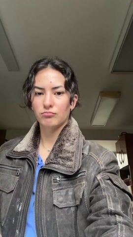 The video shows a person with short, curly brown hair wearing a brown leather jacket with a fur collar and a blue top. They are indoors, standing in front of a light-colored ceiling with two fluorescent lights. A cabinet is visible in the background. The person drinks from a small green cup, then grimaces, squinting their eyes. A blue flower clip is visible in their hair after they take the drink.