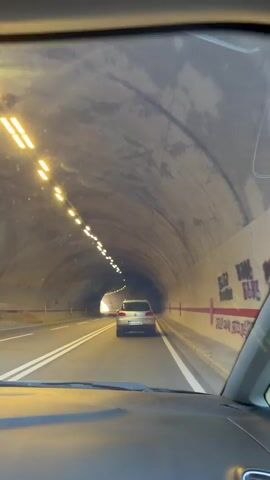 A first-person perspective video shows a car driving through a tunnel. The tunnel is illuminated by a series of lights on the ceiling. The walls of the tunnel are concrete and appear somewhat damp. There is a red stripe along the right wall. A silver car is visible ahead, also driving through the tunnel. The road has white lines marking the lanes. The video captures the motion of driving, with the view gradually changing as the car moves forward. The light at the end of the tunnel becomes brighter, indicating the car is approaching the exit.